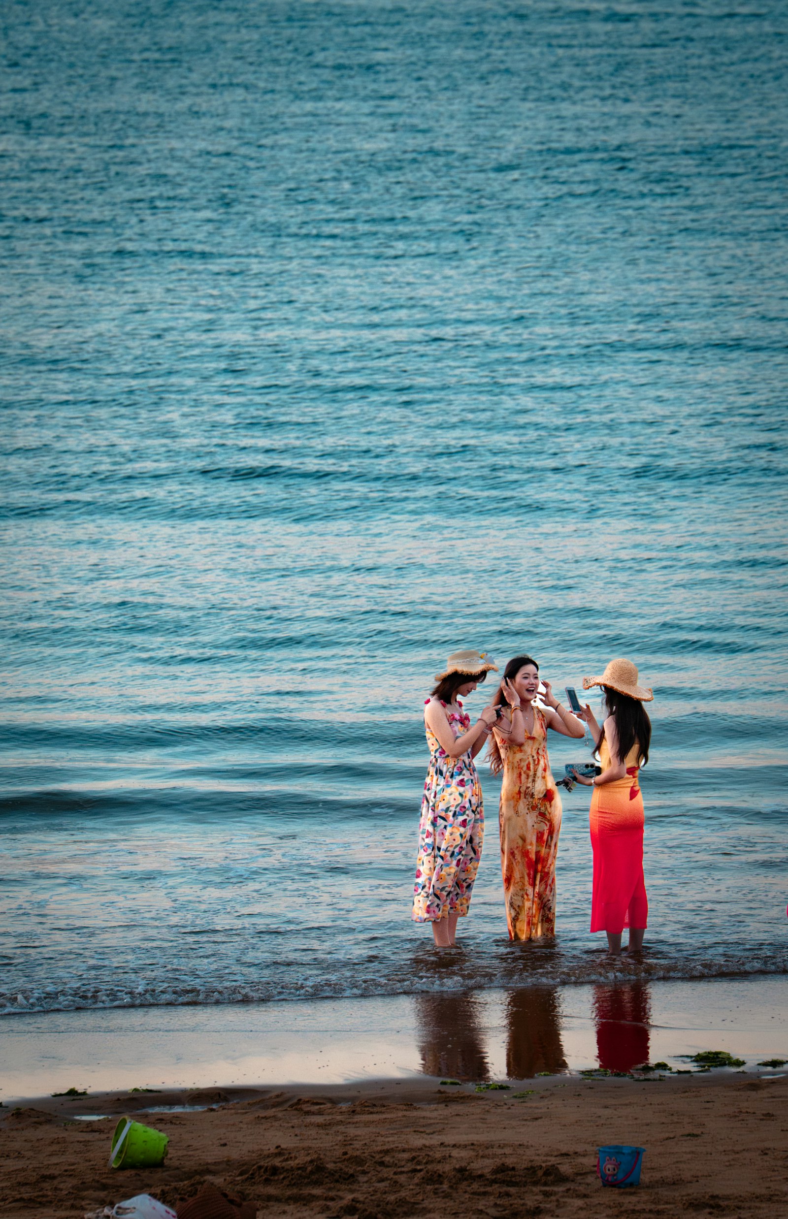 Three smiling women in bright summer dresses and straw hats standing ankle-deep in turquoise sea at the water’s edge